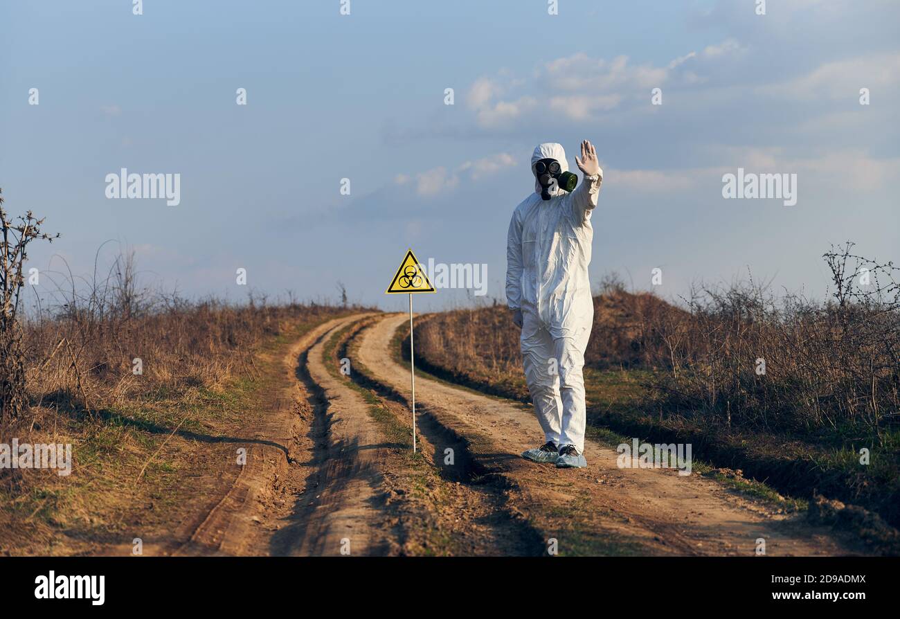 Mann in weißem Overall, Gasmaske, Handschuhe stehen auf der Straße im Feld neben dem Warnsymbol mit Stoppschild, an einem sonnigen Tag. Biohazard Zeichen Warnung vor schädlichen biologischen Substanzen und Gefahr Stockfoto