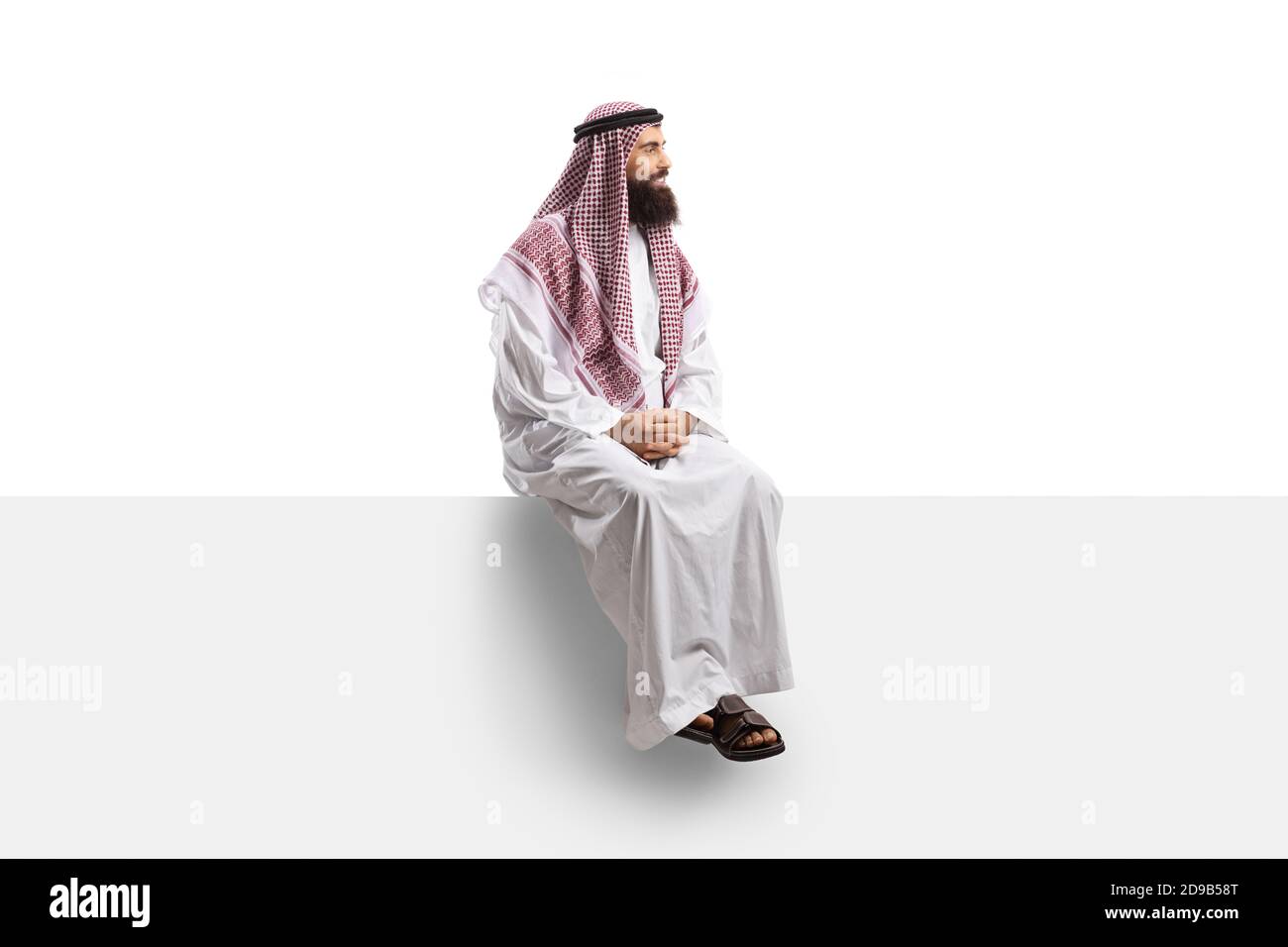 Saudi-Mann in einem Thobe sitzt auf einer leeren Tafel Und Blick zur Seite isoliert auf weißem Hintergrund Stockfoto