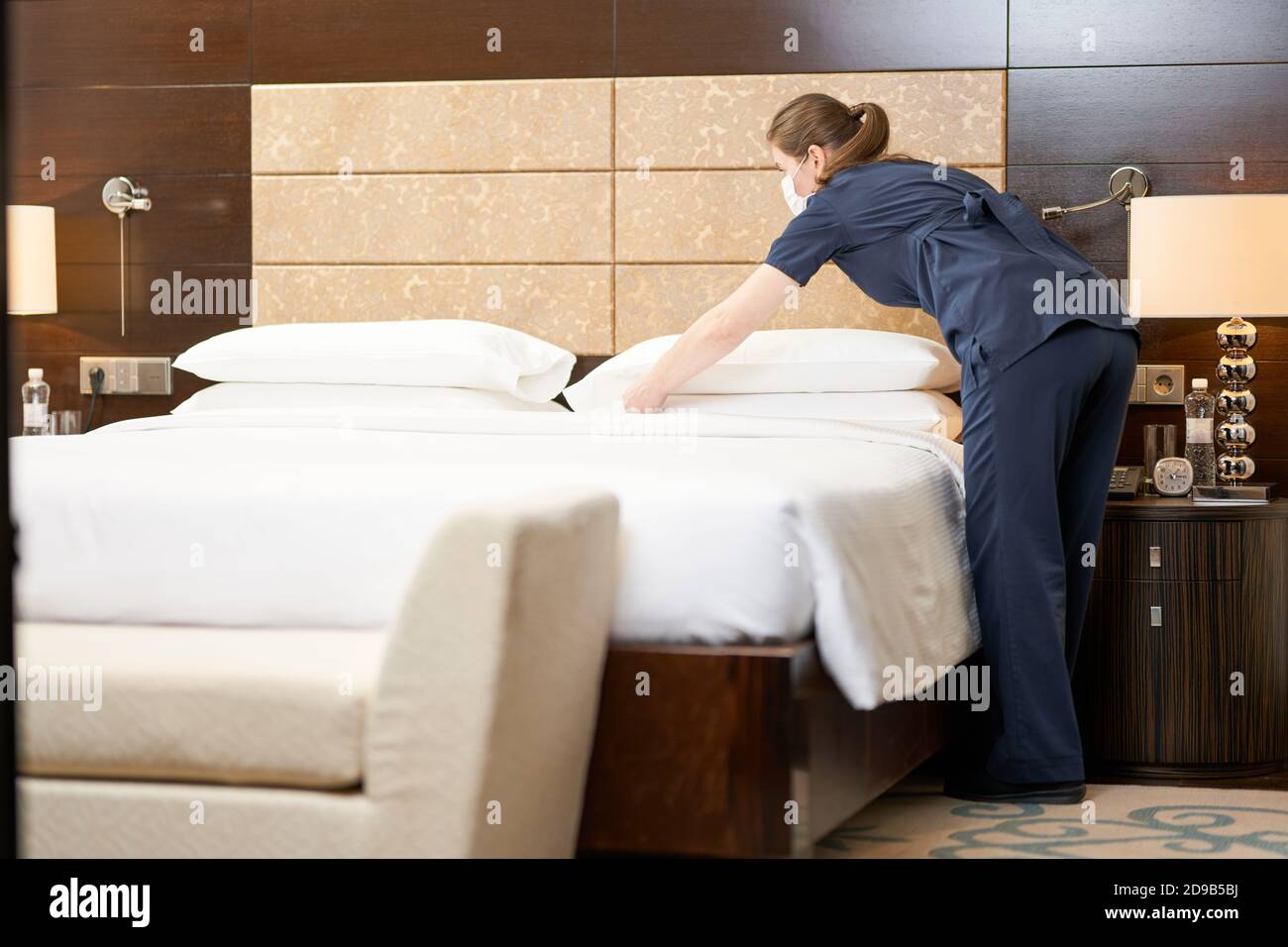 In voller Länge Foto von Maid in Maske setzen saubere Kissen Auf dem Bett im Hotelzimmer Hotelservicekonzept Stockfoto