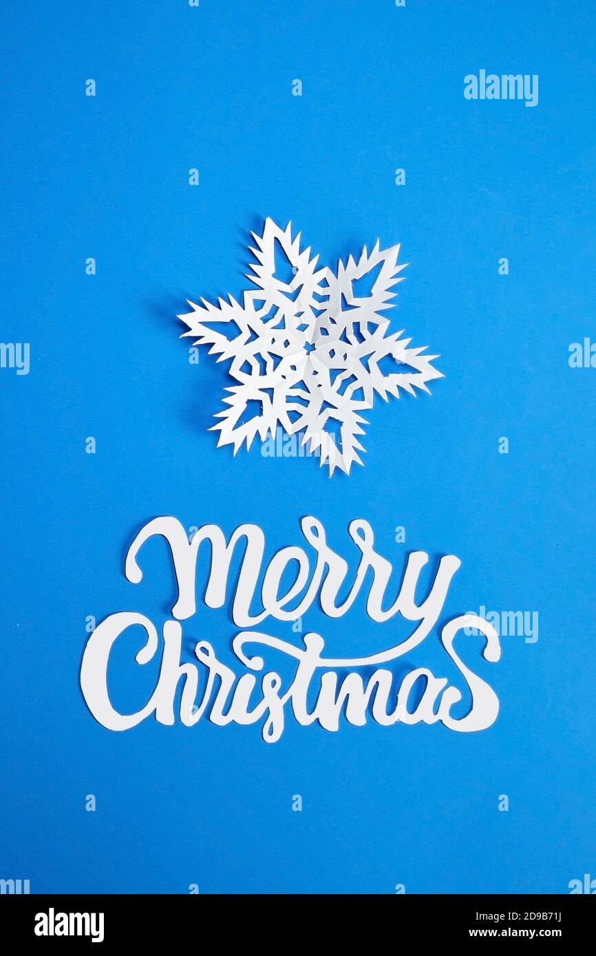 Frohe Weihnachten Text mit weißer Schneeflocke auf blauem Papier Hintergrund. Weihnachtsfeiertage Grußkarte Design. Stockfoto