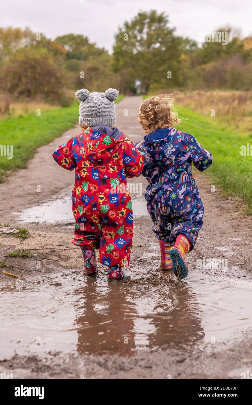 Zwei kleine Kinder, die in Gummistiefeln durch eine Pfütze gehen Und Spritzanzüge Stockfoto