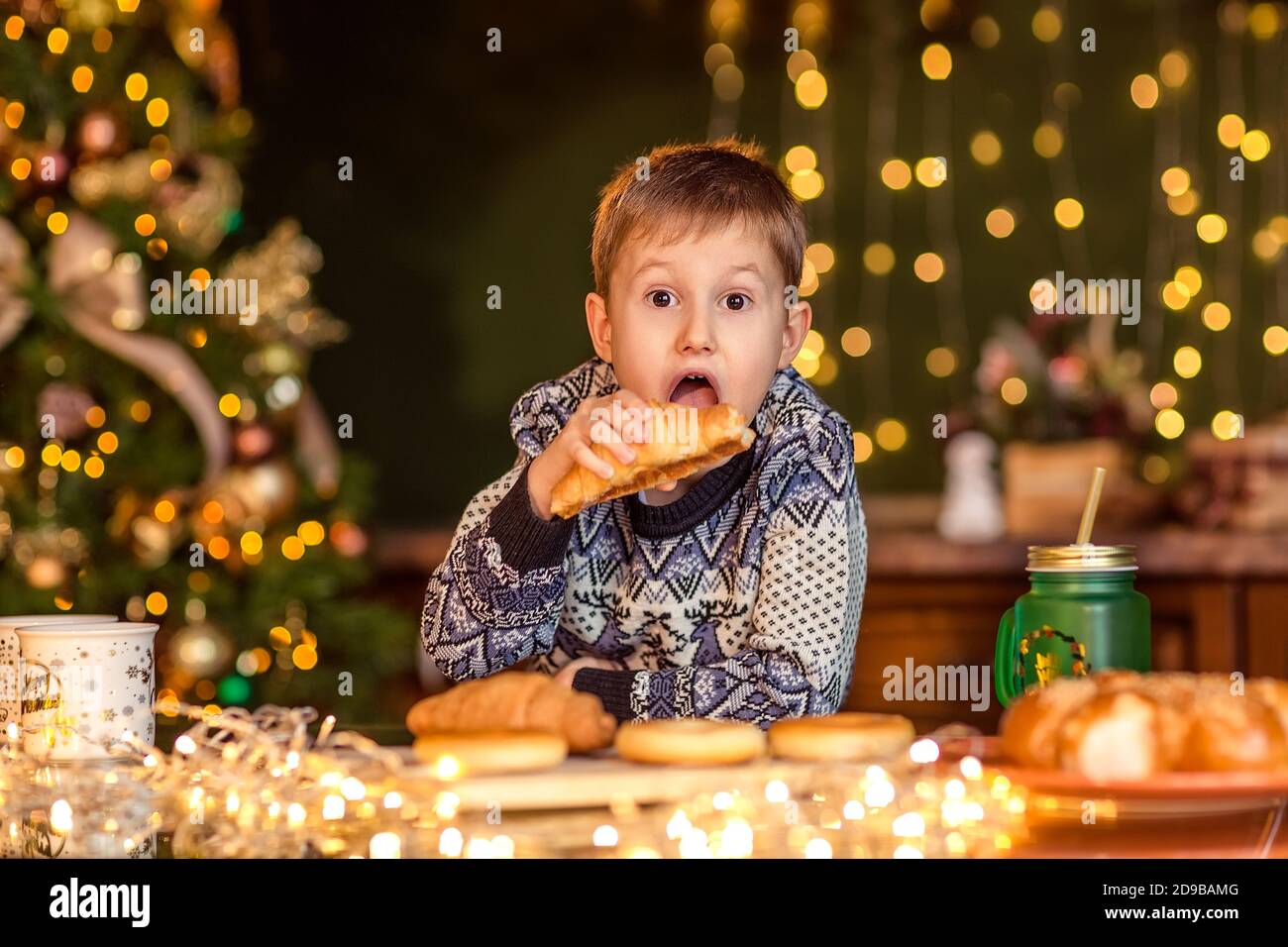 Ein Junge sitzt an einem Tisch in einer dekorierten Weihnachtsküche. Er schaut auf das Croissant und will es essen. Gemütliche Winterabende zu Hause. Konzept von Christus Stockfoto