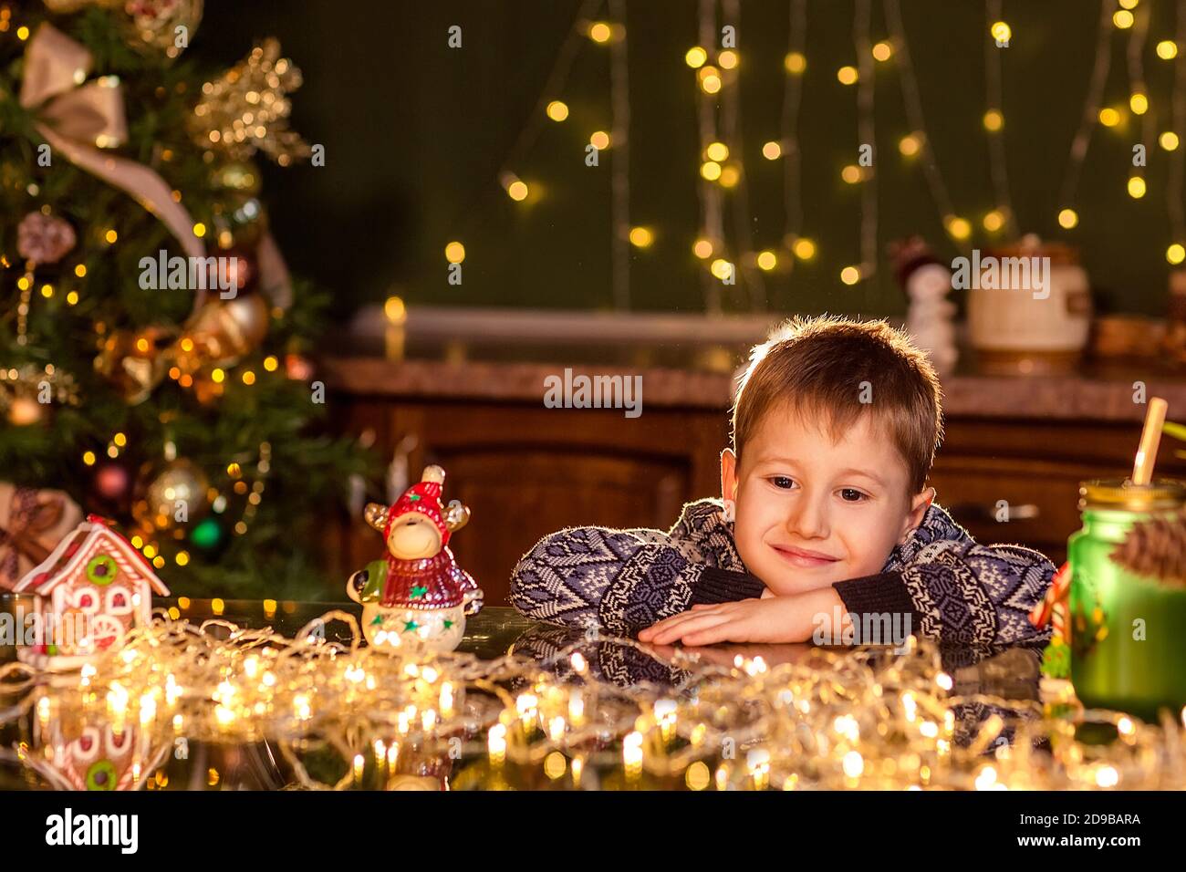 Ein Junge sitzt an einem Tisch in einer dekorierten Weihnachtsküche. Ein Spielzeug halten, um einen Weihnachtsbaum zu schmücken. Gemütliche Winterabende zu Hause. Konzept von Christus Stockfoto