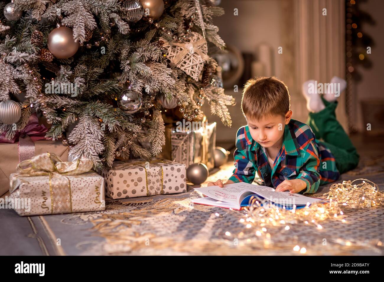 Der Junge liegt auf dem Boden neben einem geschmückten Weihnachtsbaum. Ein Buch mit Weihnachtsgeschichten lesen. Gemütliche Winterabende zu Hause. Konzept von Christm Stockfoto