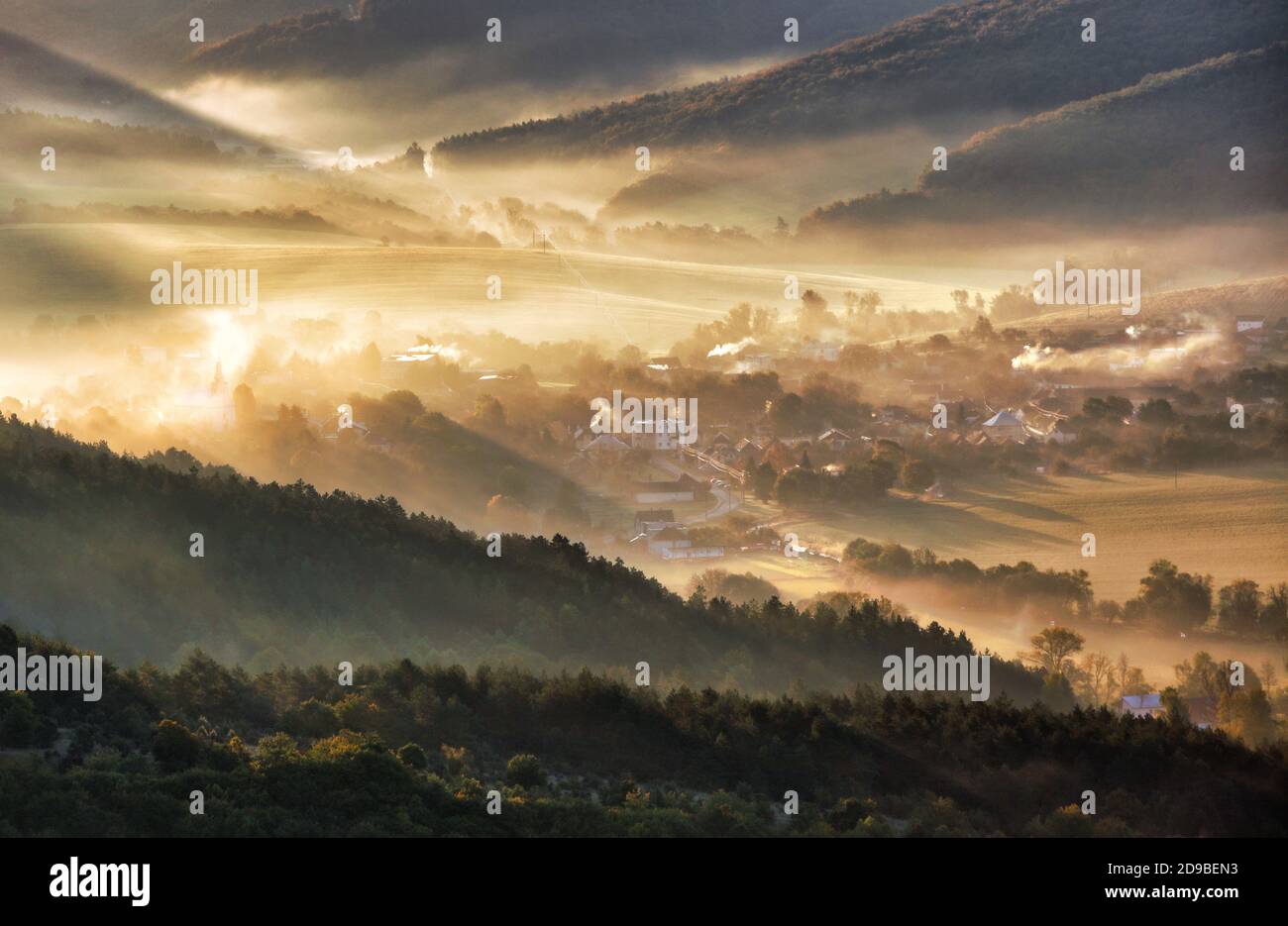 Luftaufnahme des Dorfes im Nebel mit goldenen Sonnenstrahlen bei Sonnenaufgang im Herbst. Schöne ländliche Landschaft mit Straße, Gebäuden, nebligen bunten Bäumen. Slovaki Stockfoto