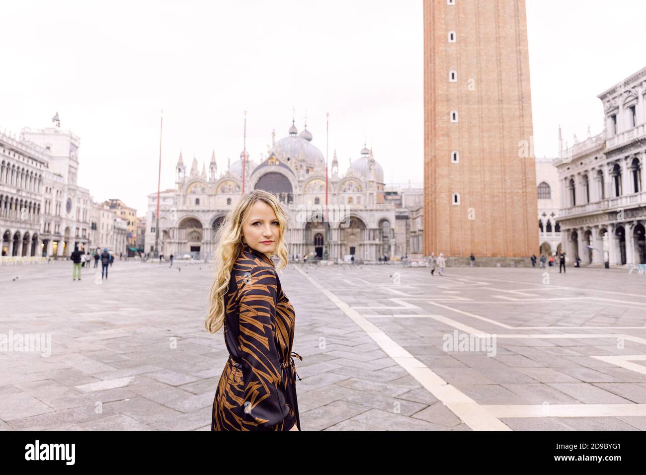 Blonde Frau, die die Kamera anschaut, in einem langen dunklen Kleid, der auf dem Markusplatz, Venedig, Italien, läuft Stockfoto