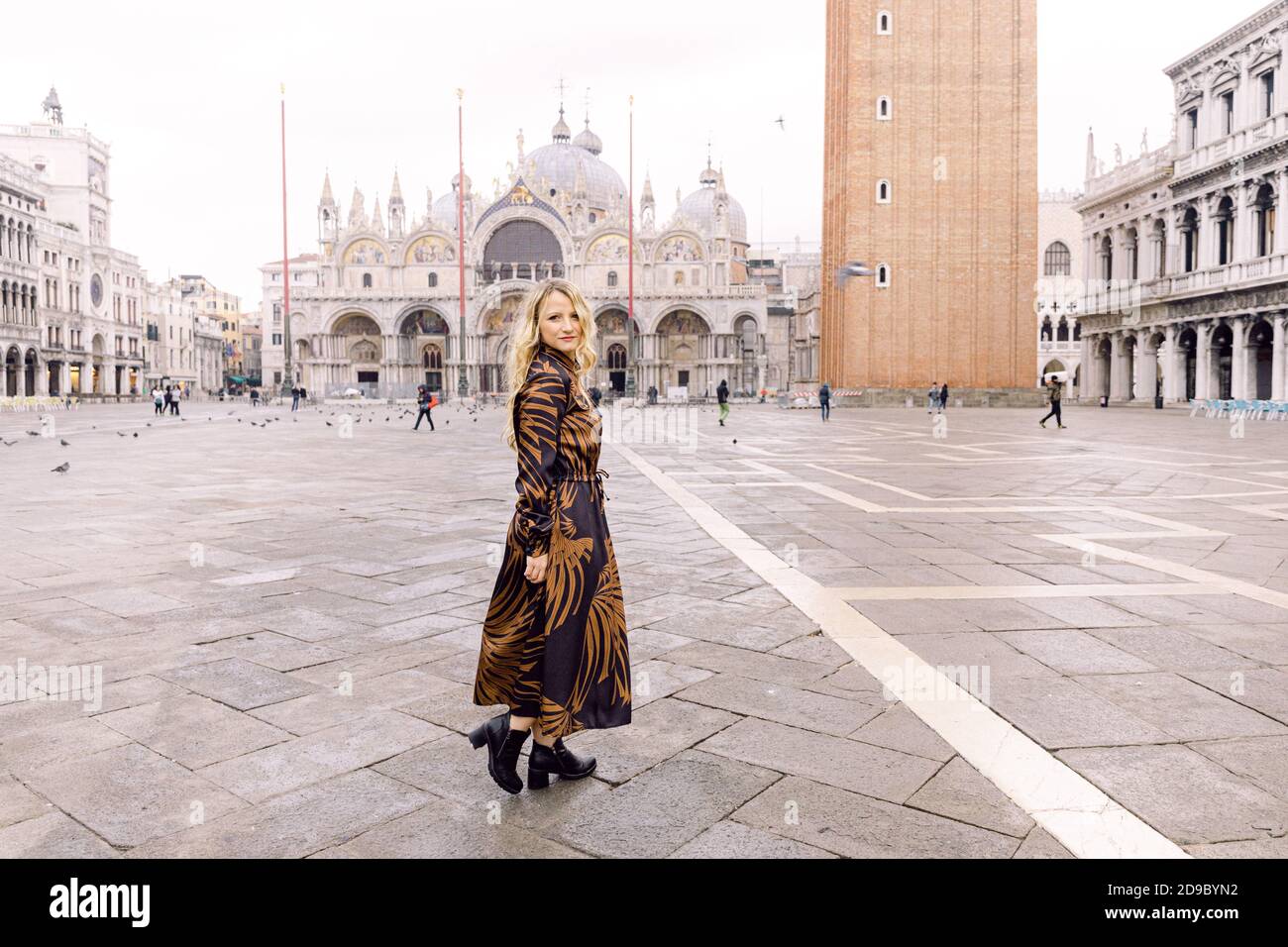 Blonde Frau, die die Kamera anschaut, in einem langen dunklen Kleid, der auf dem Markusplatz, Venedig, Italien, läuft Stockfoto