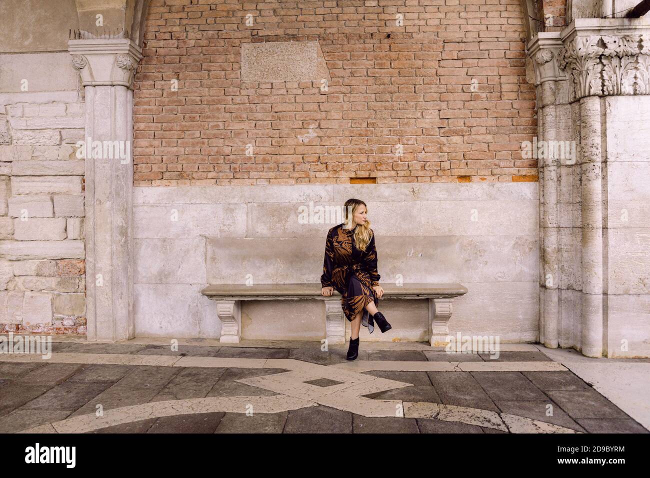 Schönes Mädchen mit einem braunen und schwarzen langen Kleid, sitzt auf einer Bank und schaut zur Seite, in Venedig, Italien Stockfoto