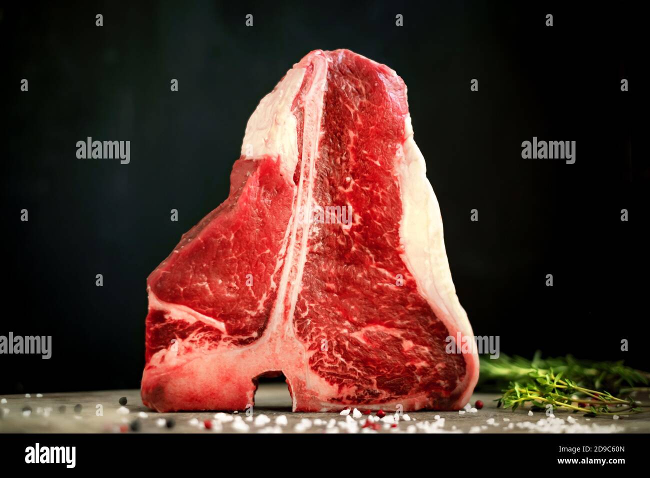 Rohes t bone Steak ist auf einem Holzbrett auf schwarzem Hintergrund, neben Salz, Pfeffer und Kräutern Stockfoto