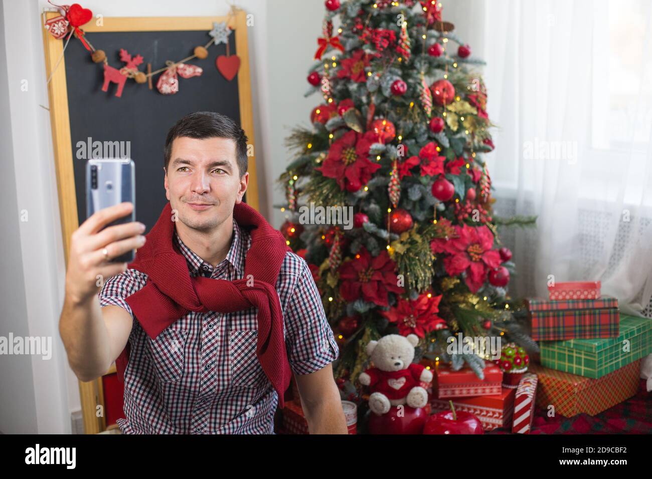 Junger Mann in der Nähe von Weihnachtsbaum zu Hause in bequemer Kleidung winkt seine Hand als Zeichen von Hallo / Auf Wiedersehen vor dem Smartphone-Bildschirm. Herzlichen Glückwunsch Stockfoto