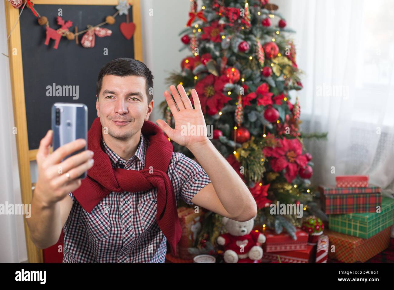 Junger Mann in der Nähe von Weihnachtsbaum zu Hause in bequemer Kleidung winkt seine Hand als Zeichen von Hallo / Auf Wiedersehen vor dem Smartphone-Bildschirm. Herzlichen Glückwunsch Stockfoto