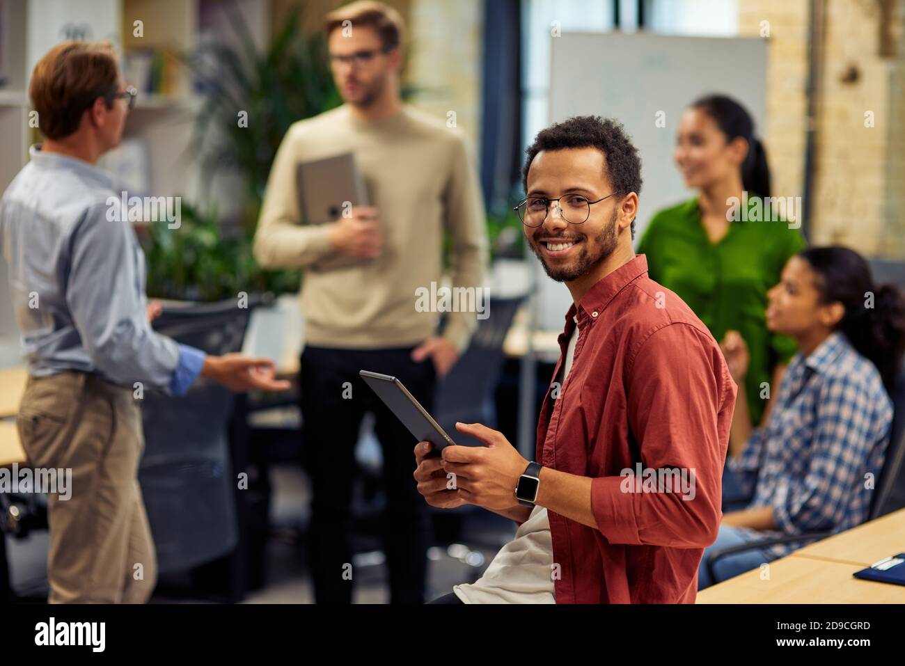 Junge fröhliche gemischte Rennen Mann hält digitale Tablette und lächelt an der Kamera während der Arbeit mit Kollegen im modernen Büro, selektive Fokus. Geschäftsleute bei der Arbeit Stockfoto
