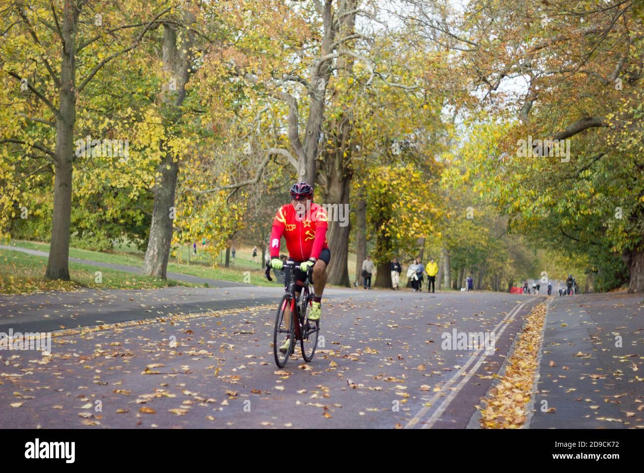 Begeisterter Radfahrer in rot oben Reiten bergauf Straße mit herbstlichen Blättern in Greenwich Park bedeckt. London, England, Vereinigtes Königreich Stockfoto