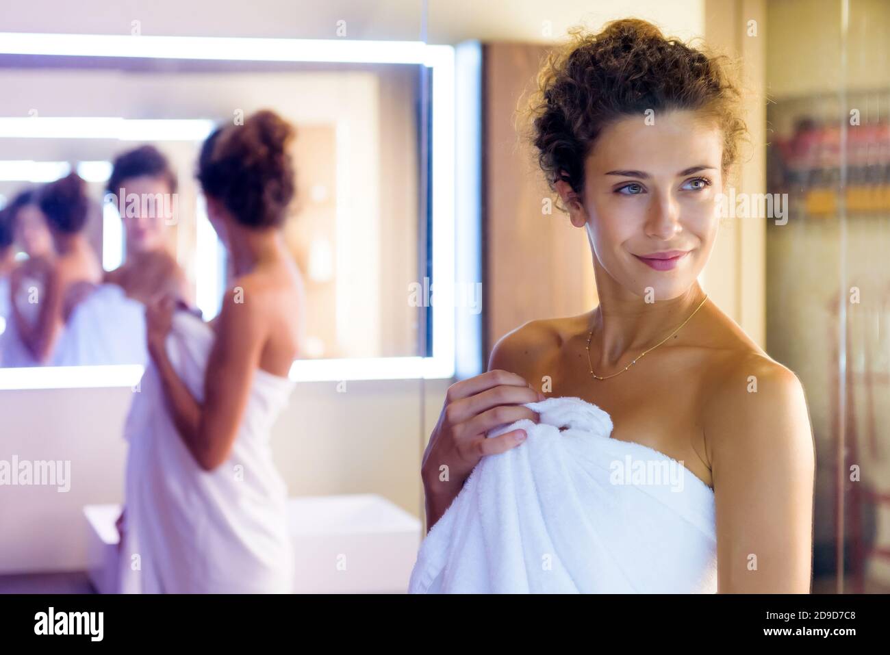 Lächelnde Frau reflektierte mehrmals in einem Spiegel, als sie Posen in einem sauberen weißen Handtuch im Badezimmer vorbereiten Für eine Dusche Stockfoto