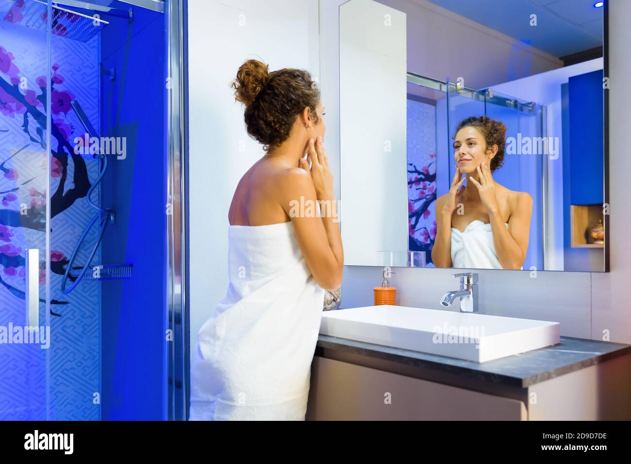 Erfrischte junge Frau in einem sauberen weißen Handtuch suchen gewickelt Bei sich selbst in einem Badezimmerspiegel nach dem Duschen in einem Hygiene- oder Schönheitskonzept Stockfoto