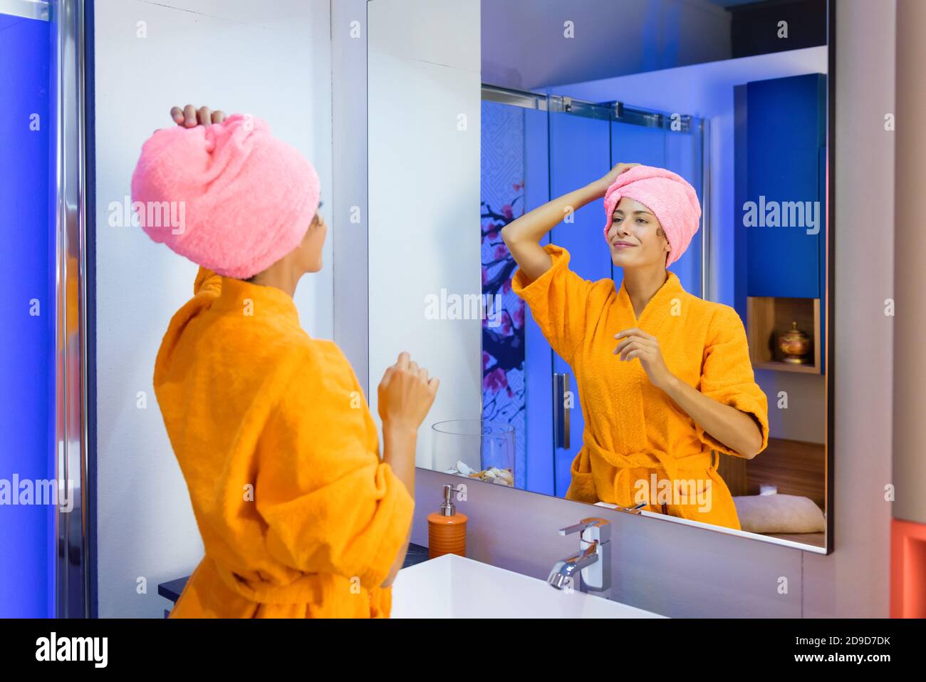 Junge Frau trägt einen leuchtend orangefarbenen Bademantel mit ihr Haare in einem Handtuch nach dem Duschen Blick auf sich in Ein Badezimmerspiegel mit einem ruhigen s Stockfoto