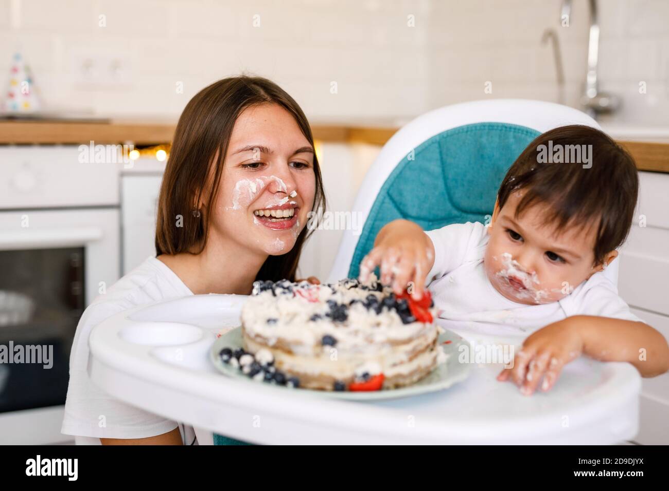 Glückliche Mutter mit einem Jahr alten Baby feiert mit ersten Geburtstag mit weißer Küche auf Hintergrund. Stockfoto