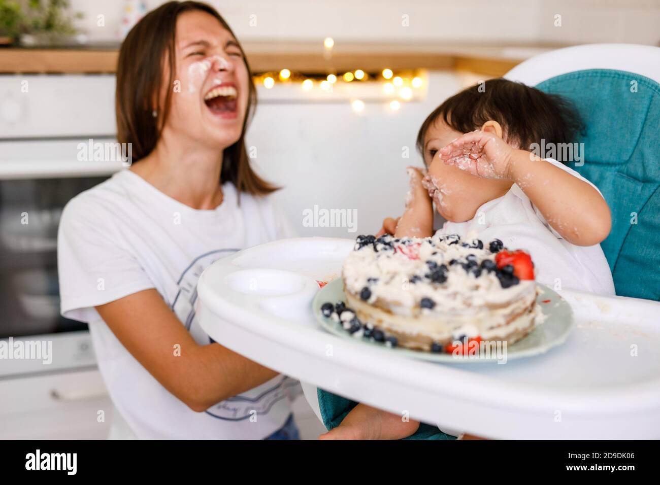 Glückliche Mutter mit einem Jahr alten Baby feiert mit ersten Geburtstag mit weißer Küche auf Hintergrund. Stockfoto