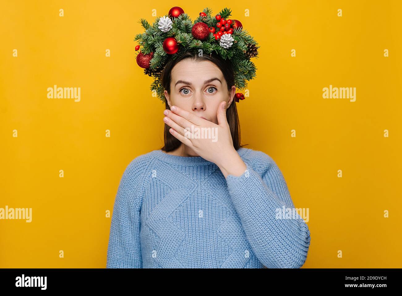 Schockiert überrascht junge Frau in traditionellen Weihnachtskranz bedeckt Mund mit Händen, tragen gemütliche blaue Pullover, isoliert auf gelbem Hintergrund Stockfoto
