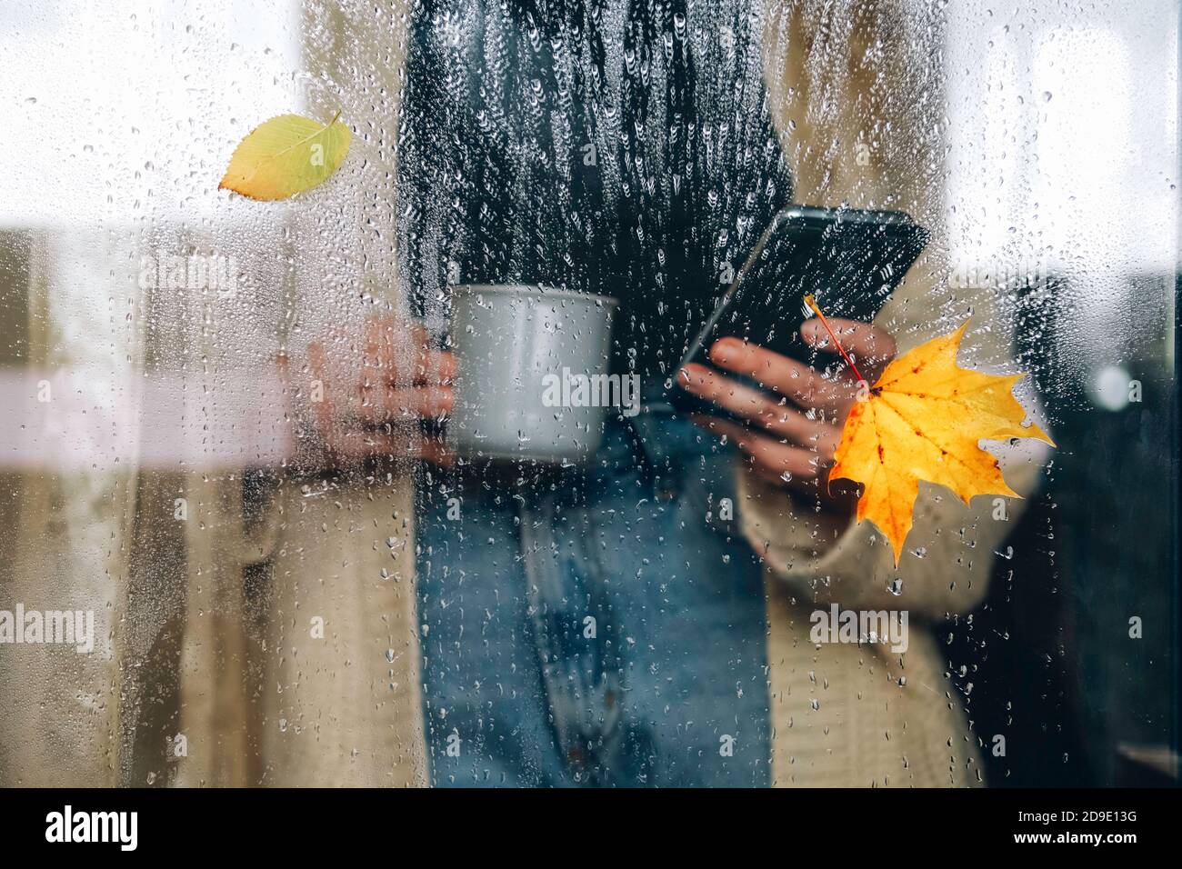 Frau in warmer Strickjacke mit Handy und Tasse Tee an einem regnerischen Herbsttag am Fenster stehend. Stockfoto