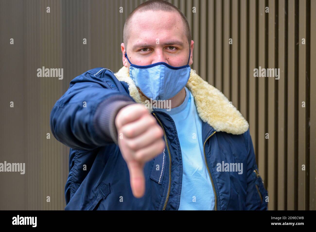 Junger Mann trägt ein wiederverwendbares Tuch Gesichtsmaske während der Covid-19 Pandemie zeigt dies mit Daumen nach unten Alles ist nicht gut oder um seine auszudrücken Stockfoto