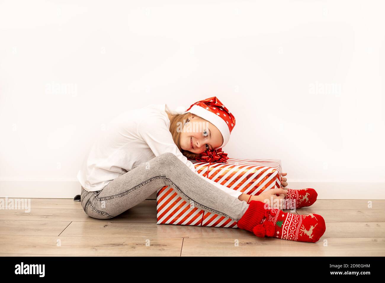 Ein schönes Mädchen in legerer Kleidung mit Weihnachtszubehör sitzt auf dem Boden und umarmt eine große Geschenkbox mit einem roten Band und einer Schleife. In voller Länge Stockfoto