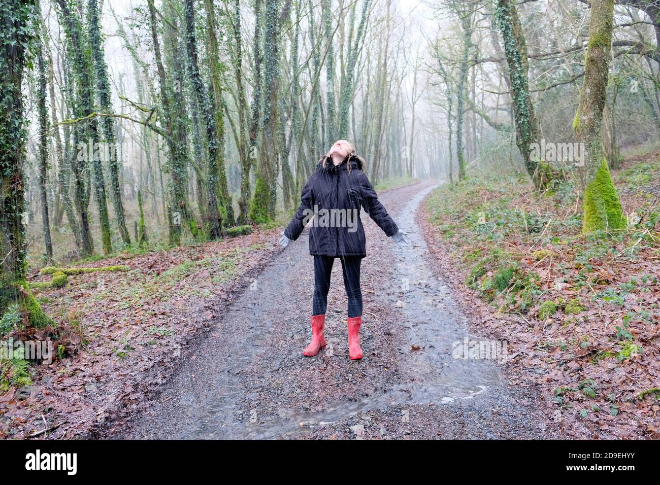 Junge Frau 20s im Wintermantel Gummistiefel stehen am Regentag und schauen mit Freude auf Regentropfen, die auf dem Land in Wales UK KATHY DEWITT fallen Stockfoto