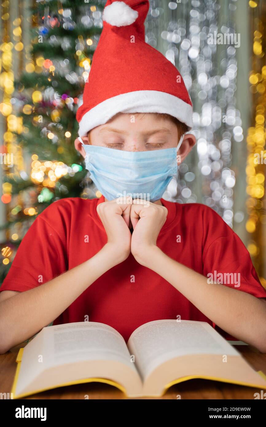 Kind mit rotem Weihnachtsmann Hut vor der Bibel beten auf Weihnachten dekoriert Hintergrund mit Geschenken auf Tisch - Konzept des Gebets für einen Weihnachtsabend. Stockfoto