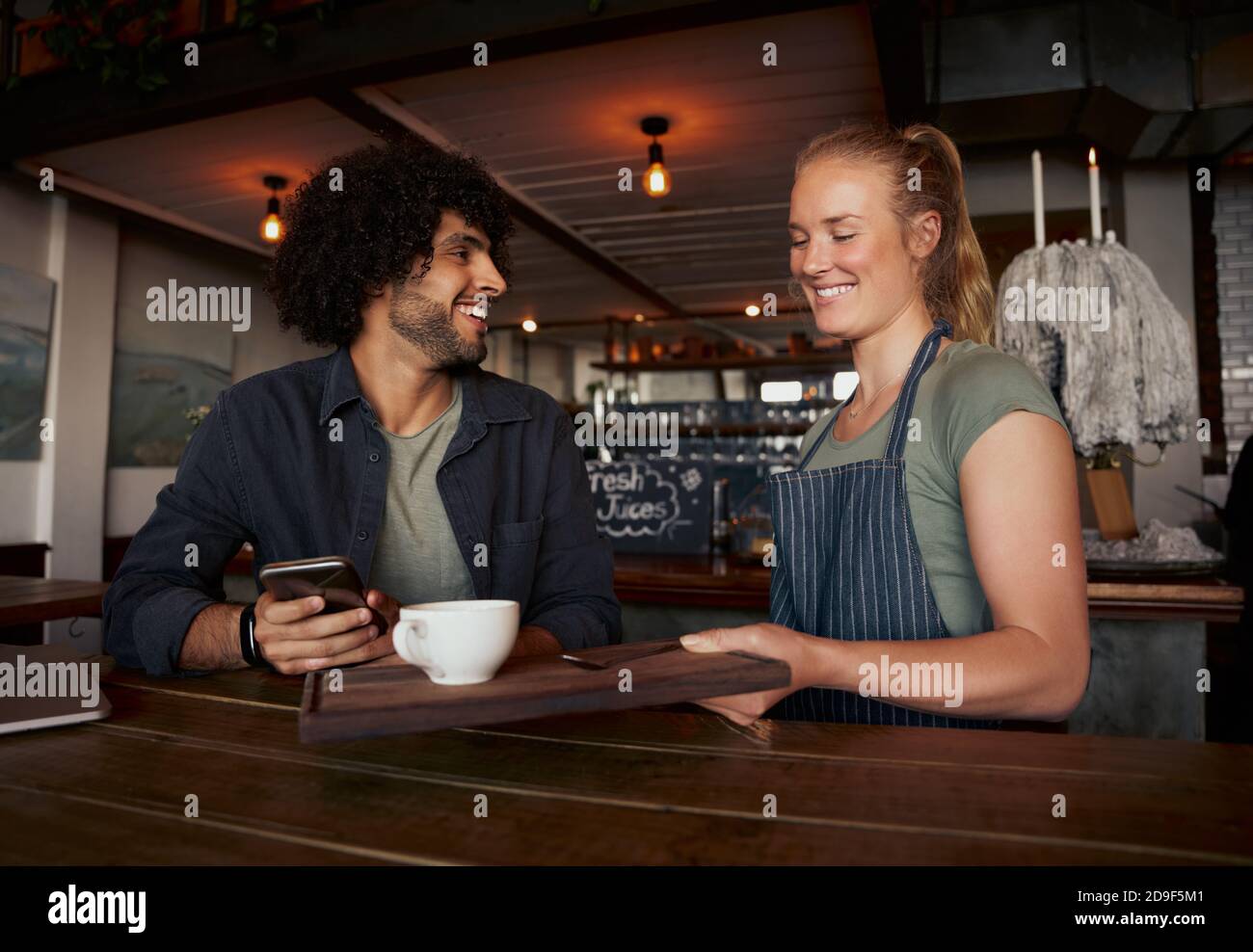 Fröhliche weibliche Kellnerin serviert Kaffee zu jungen männlichen Kunden mit Lockiges Haar mit dem Smartphone im modernen Café Stockfoto