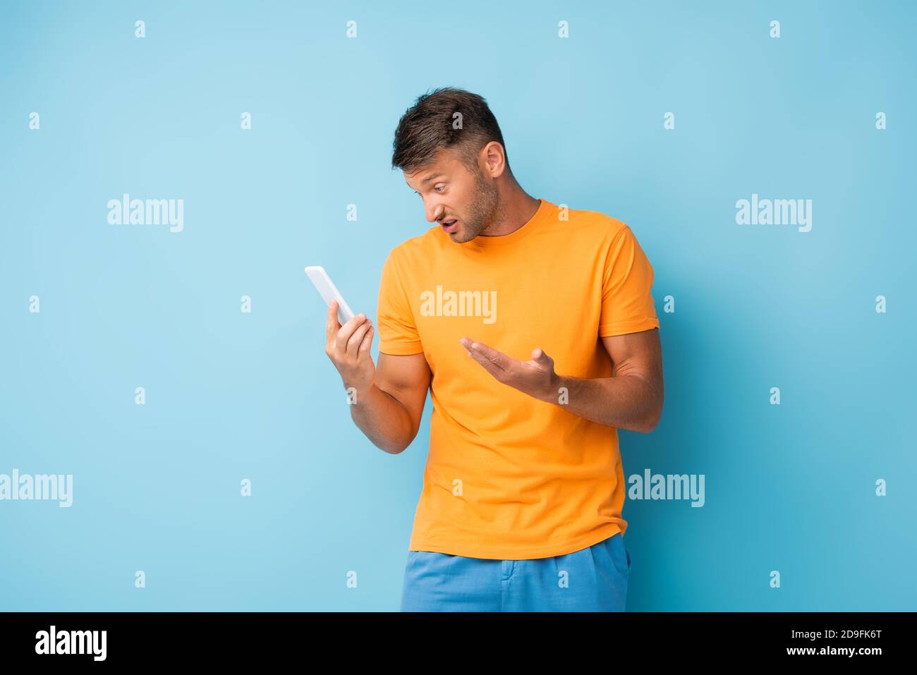 Unzufriedener Mann in T-Shirt hält Smartphone auf blau Stockfoto