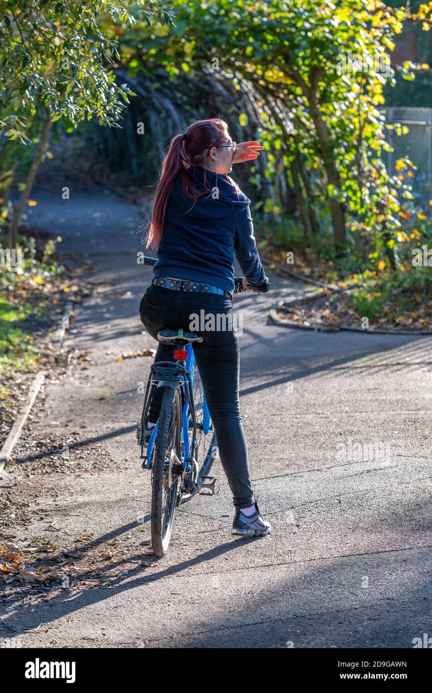 Eine Frau, die mit dem Fahrrad durch einen Park fährt und ihre Hand über die Augen hält, um sie vor der Sonne zu schützen und die Straße vor sich zu sehen. Stockfoto