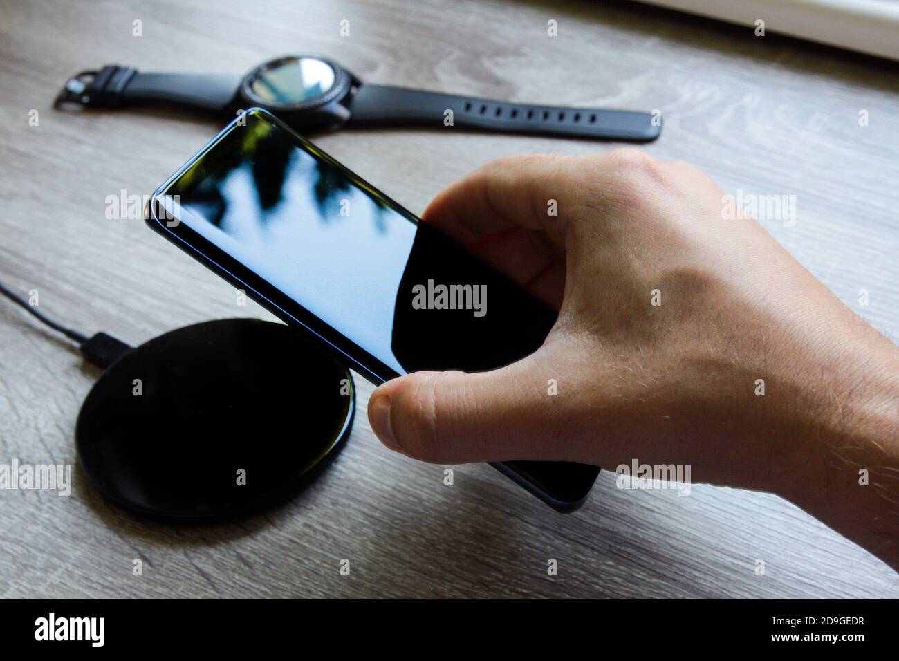 Das in der Hand gehaltene Smartphone wird auf das kabellose Ladegerät gestellt. Intelligente Uhr im Hintergrund Stockfoto