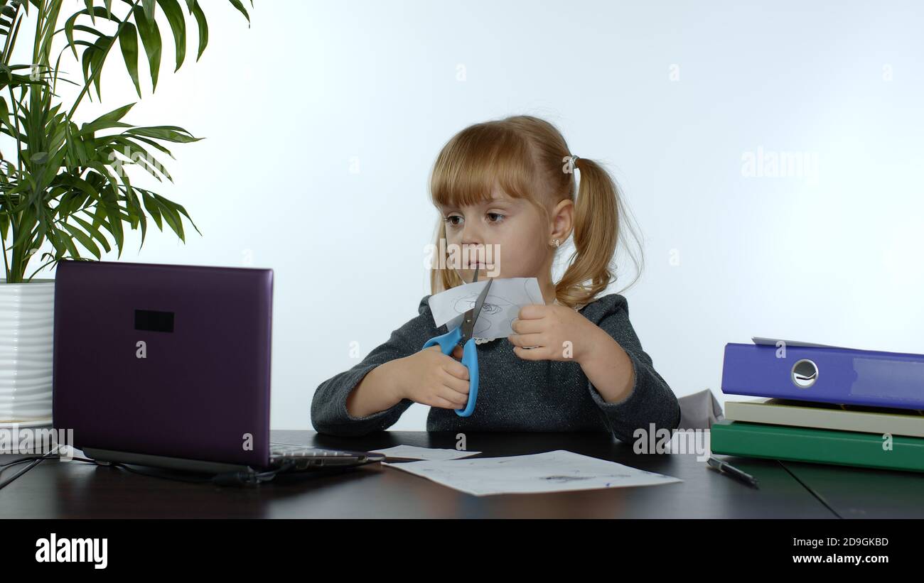 Kind Schulmädchen lernt Unterricht zu Hause sitzen am Tisch Schneiden mit Scheren Formen aus Papier. Kinder Mädchen Fernunterricht. Studieren während Coronavirus Quarantäne. Weißer Hintergrund Stockfoto