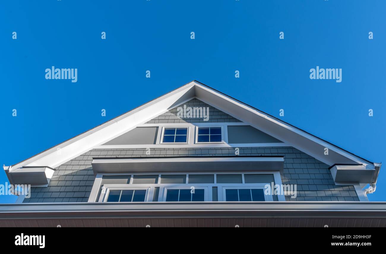 Dekorativer Giebel mit weißen Dachrinnen und Soffit, grauen Schindelschiefern, Ausstoßfenstern, eingerahmt von einem dicken weißen Rahmen mit blauem Himmel auf einer neuen Konstruktion Stockfoto