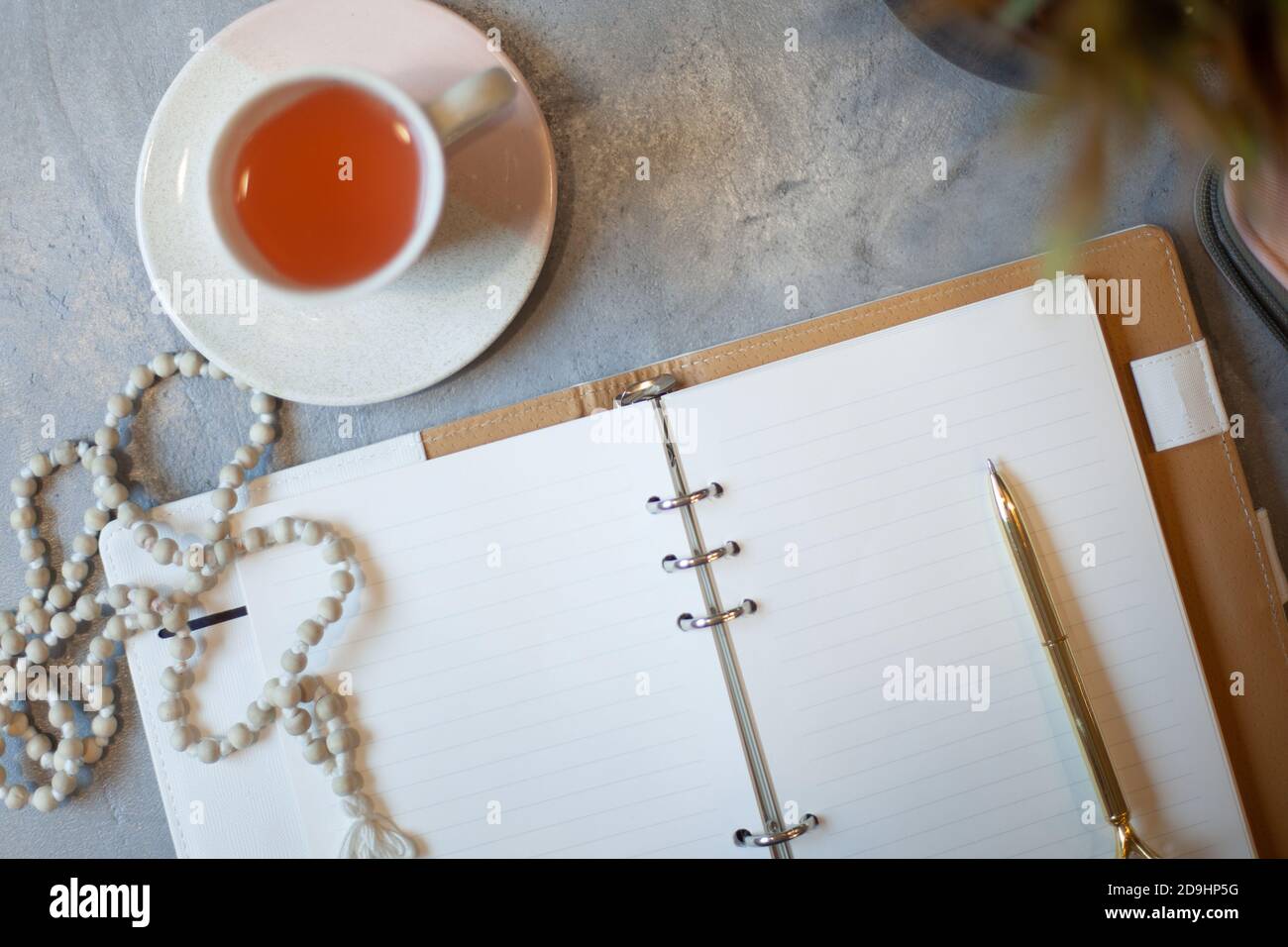 Geöffnetes Notizbuch mit sauberen Laken, goldenem Stift, Rosenkranz, pinkfarbenem Etui und einer Tasse Tee in der Nähe einer Vase Stockfoto