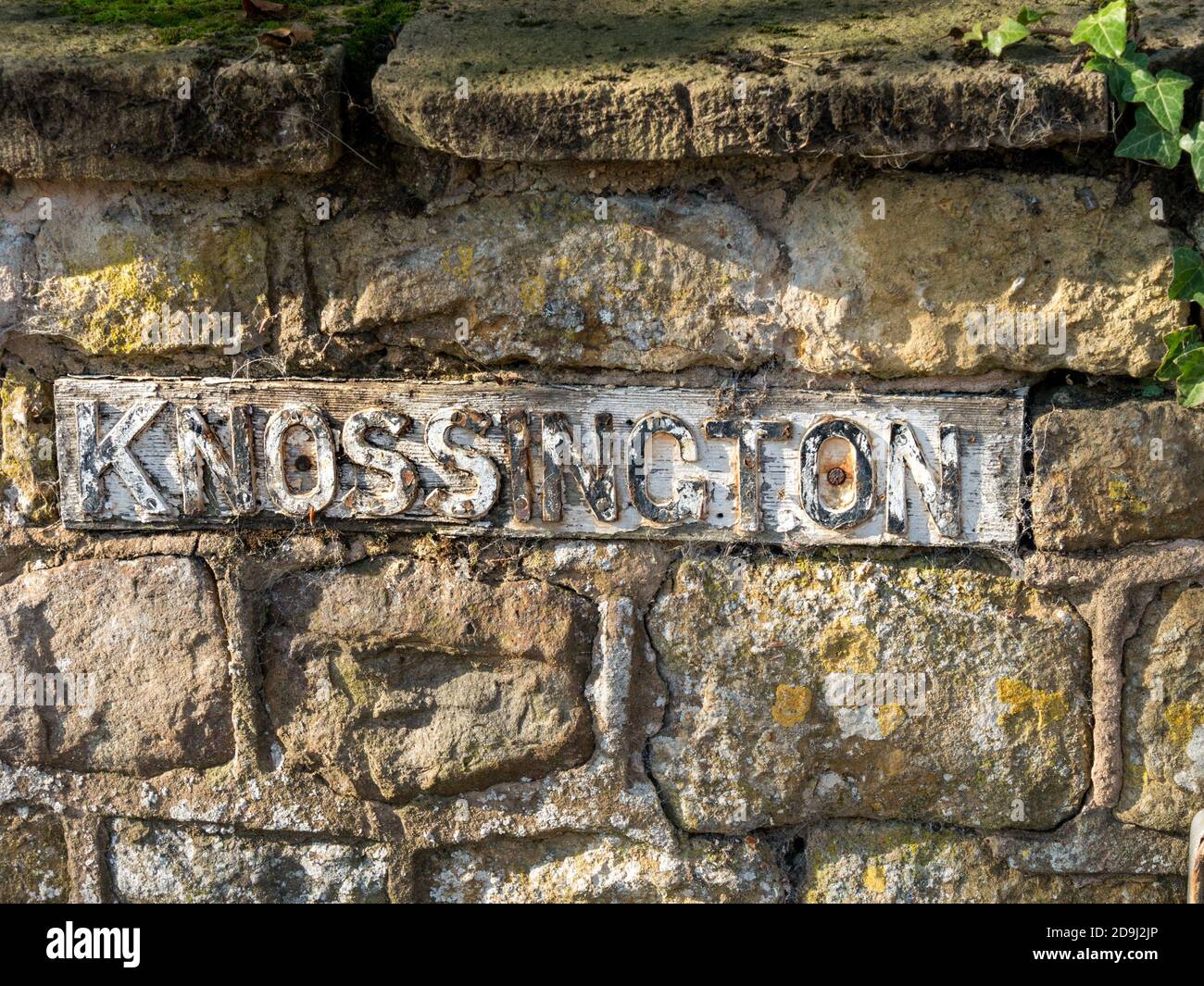 'Knossington' hübsches, altes gusseisernes Schild mit erhabenem geprägtem Schriftzug in alter Steinwand, Knossington, Leicestershire. Stockfoto
