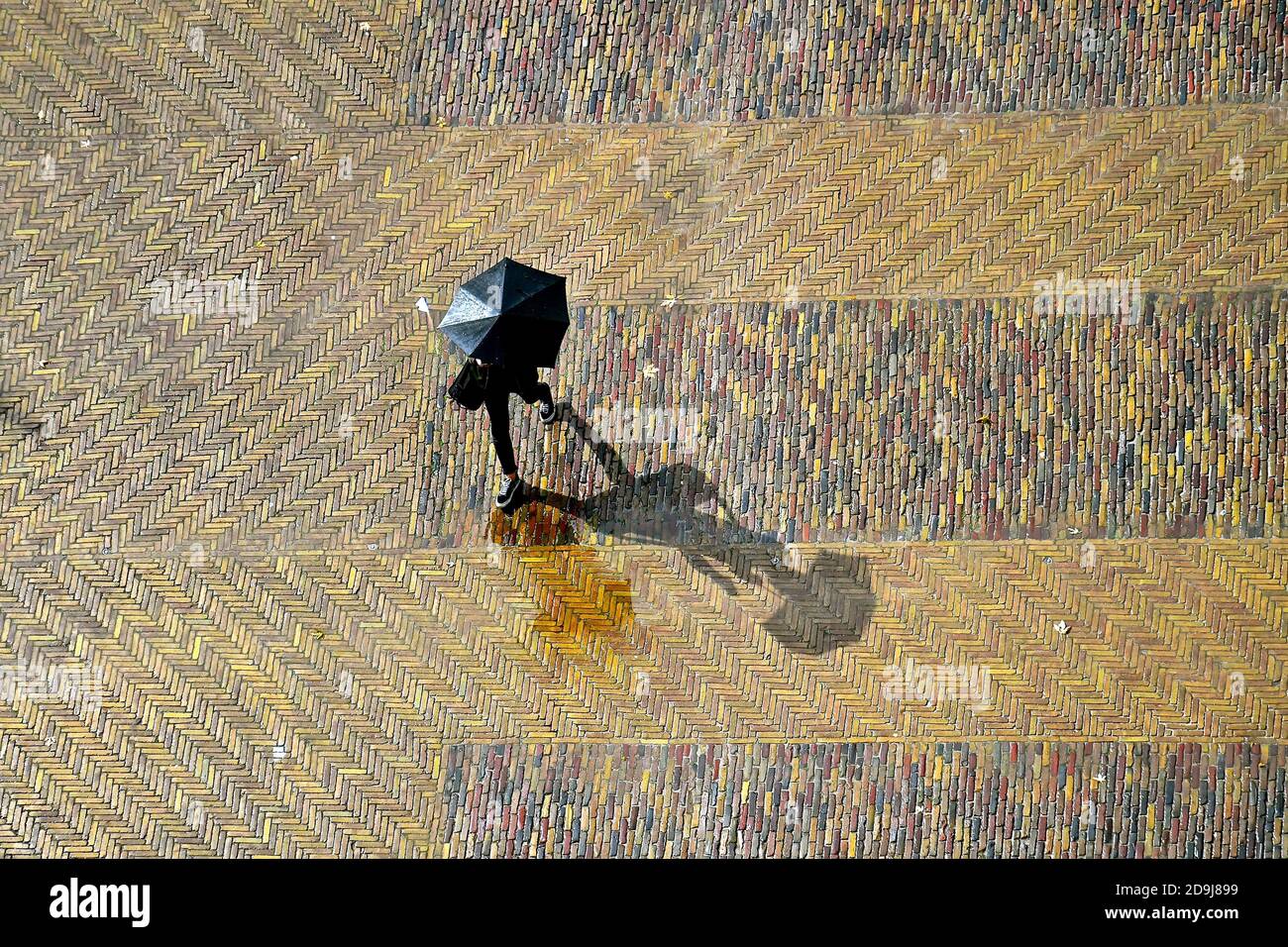 Luftaufnahme eines Fußgängers, der über einen alten, gepflasterten Marktplatz unter einem schwarzen Regenschirm bei Regen und Sonnenschein mit einem langen Schatten geht. Stockfoto