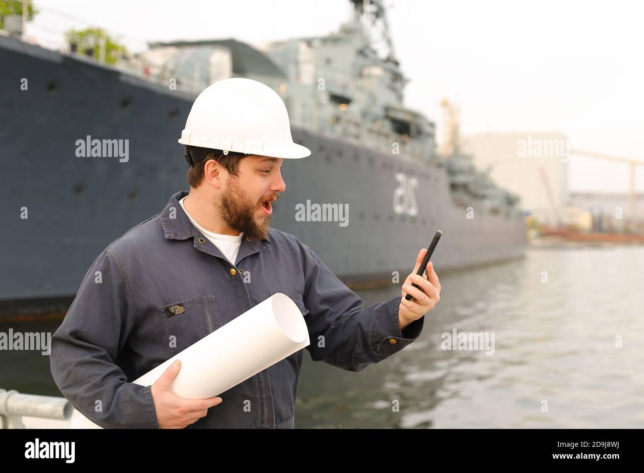 Marineingenieur im Helm hält VHF Walkie Talkie und Papiere in der Nähe des Schiffes im Hintergrund. Stockfoto