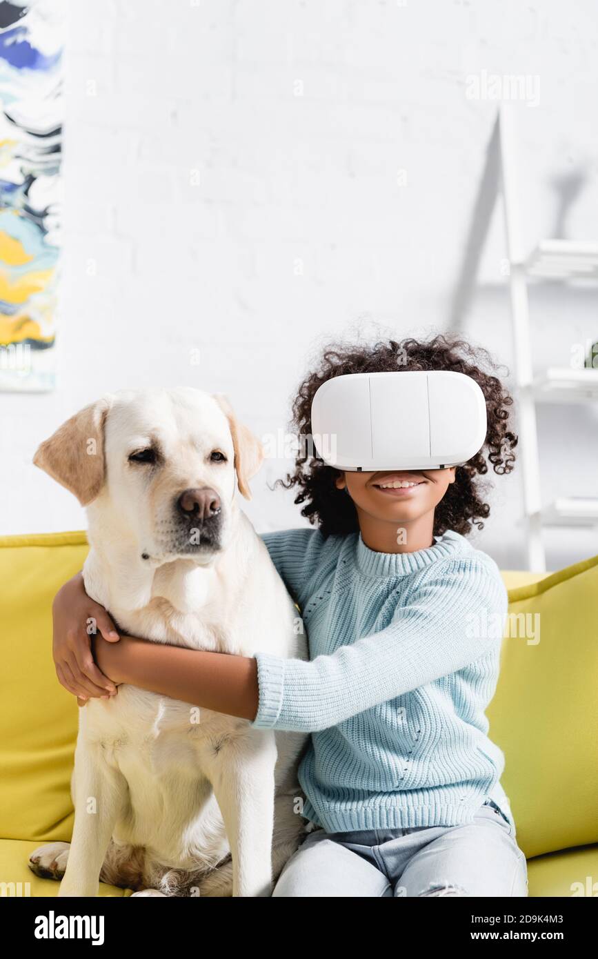 Glücklich lockig afroamerikanische Mädchen umarmt Retriever, während das Tragen vr Headset, sitzt auf dem Sofa auf verschwommenem Hintergrund Stockfoto