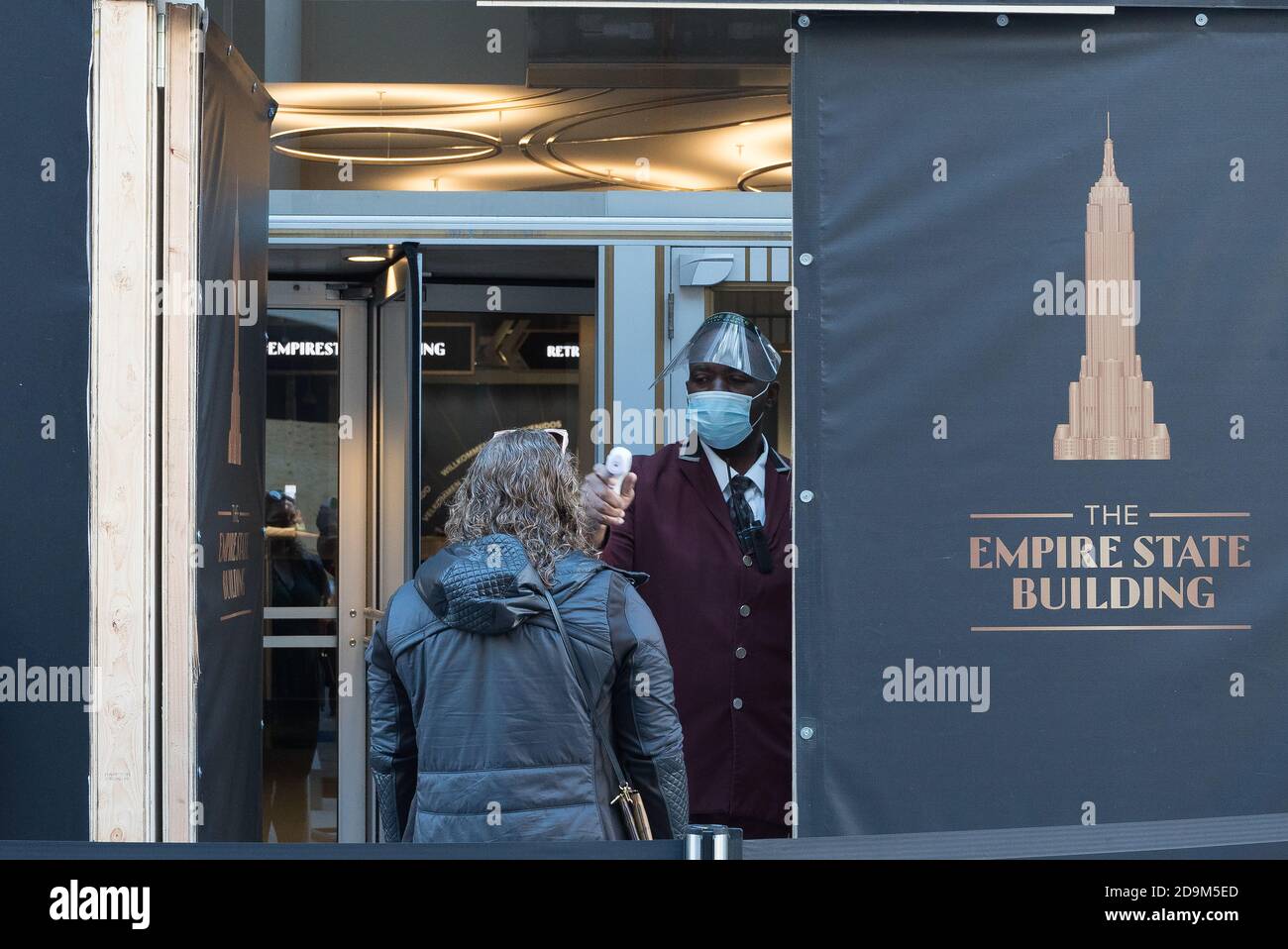 Mitarbeiter des Empire State Building, der eine Maske trägt, misst die Körpertemperatur einer Frau, bevor sie das Gebäude betreten kann. Stockfoto