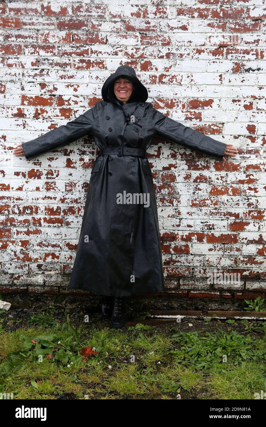 Reife weiße kaukasische Frau in Designer-Regenbekleidung, genießen den  Regen und im Freien. Glänzendes schwarzes Gummi-Regenmantel mit Sowester,  der im Regen um das Fischerdorf herumläuft, schwarzes Fell im Kontrast zum  kugeligen Boot Stockfotografie -