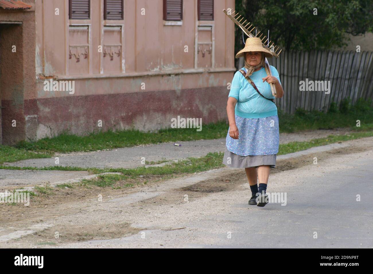 Siebenbürgen, Rumänien. Ältere Frau auf der Dorfstraße mit landwirtschaftlichen Werkzeugen. Stockfoto