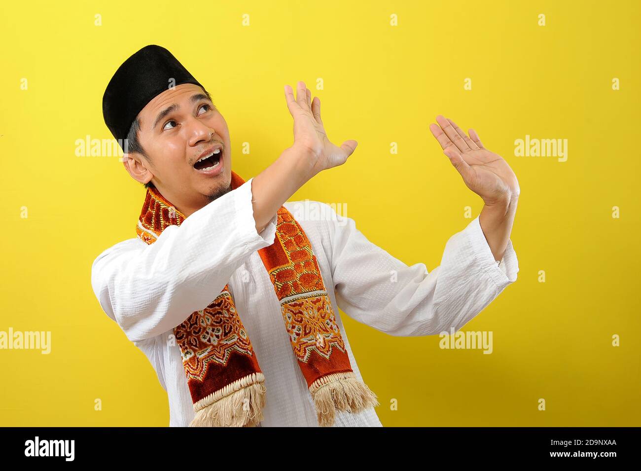 Ein bärtiger junger asiatischer Muslim, der sich mit ausgestreckten Händen vor etwas schützt, das von oben zu ihm kommt, isoliert auf gelbem Hintergrund Stockfoto