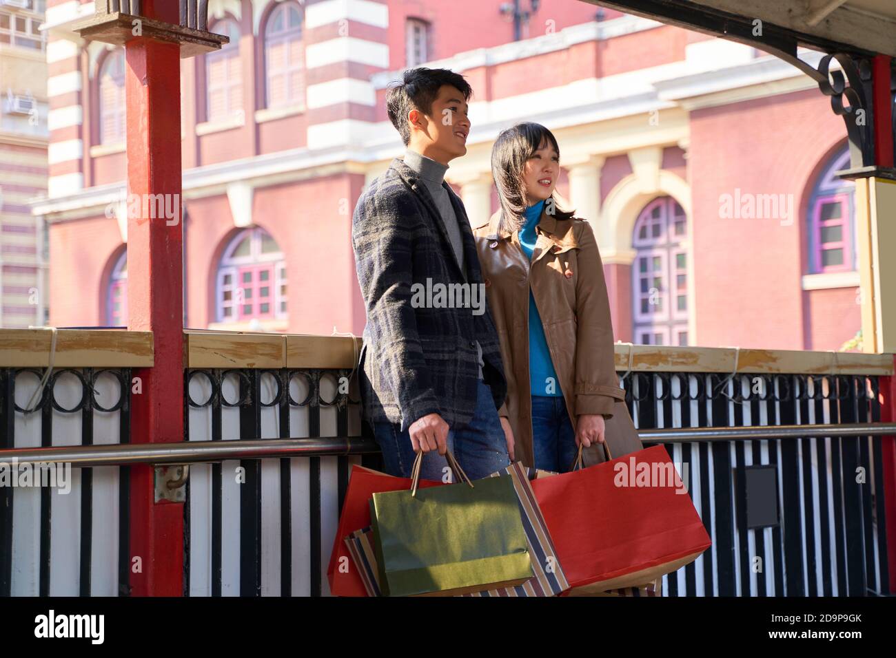 Junges asiatisches Paar, das auf einer Fußgängerüberführung steht und einkaufen hält Taschen Stockfoto