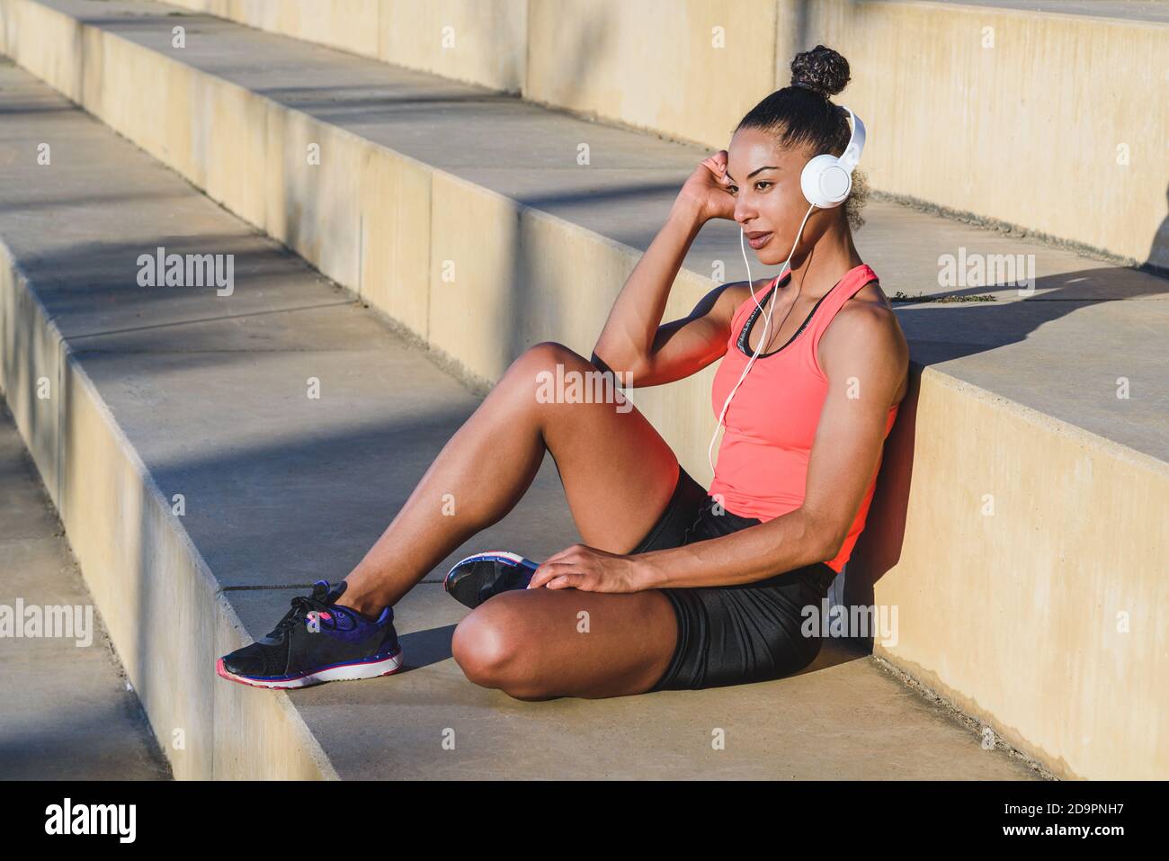 Horizontales Porträt einer attraktiven jungen afroamerikanischen Frau gekleidet in Sportbekleidung und tragen moderne weiße Kopfhörer sitzen auf einigen ruhen Schritte Stockfoto