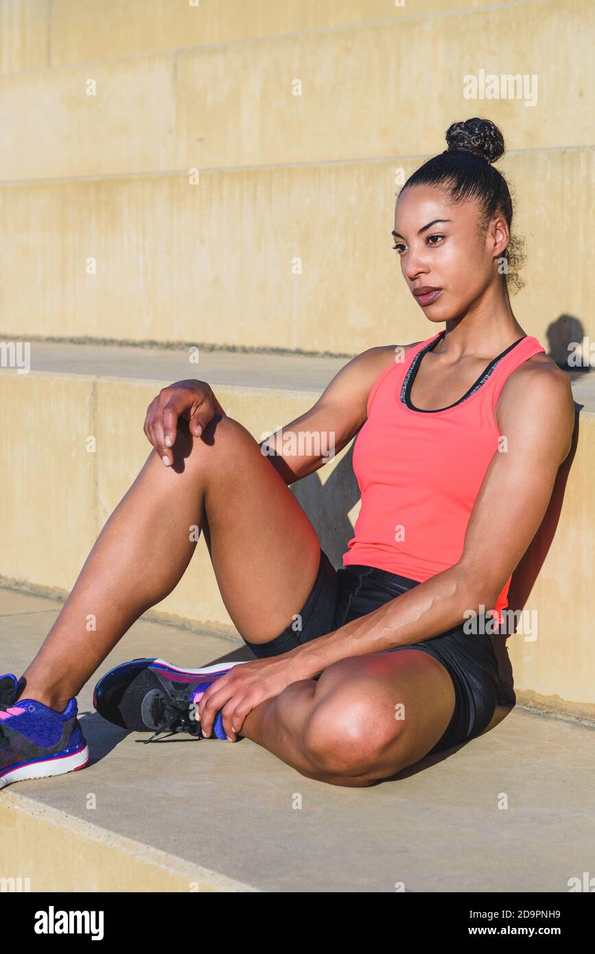 Vertikale Porträt von jungen attraktiven afroamerikanischen Frau in gekleidet Sportswear sitzend ruht auf einigen Schritten mit selbstbewusster Ausdruck und Gießen Stockfoto