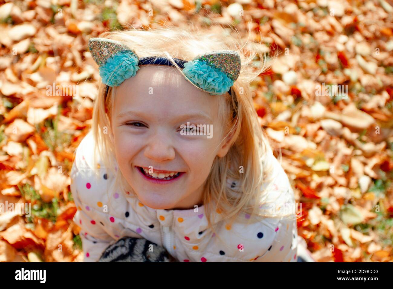 Liebenswert blonde Mädchen auf gelb gefallenen Blättern sitzen Blick auf einen schönen Herbsttag. Spielen im Freien im Park. Mädchen trägt Katze Stirnband. Stockfoto