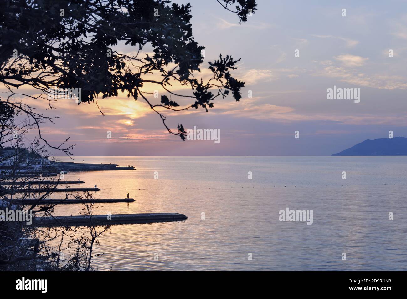 Dramatische Sonnenuntergang mit schönen Wolken und Baum auf einem Hintergrund. Piers und Berge blau gefärbt. Entspannend und ruhig ruhigen Ort. Dämmerung Zeit am Meer. Stockfoto