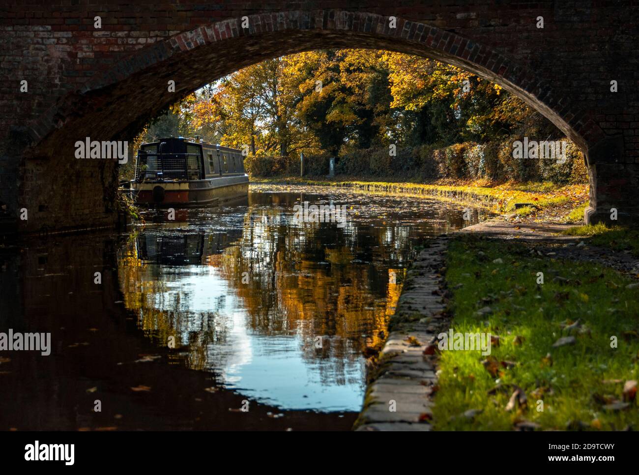 Ein Blick auf ein enges Boot unter dem Bogen einer gemauerten Fußbrücke, die sich in das stille Wasser des Kanals spiegelte, mit herbstlichen Farben in den Bäumen Stockfoto