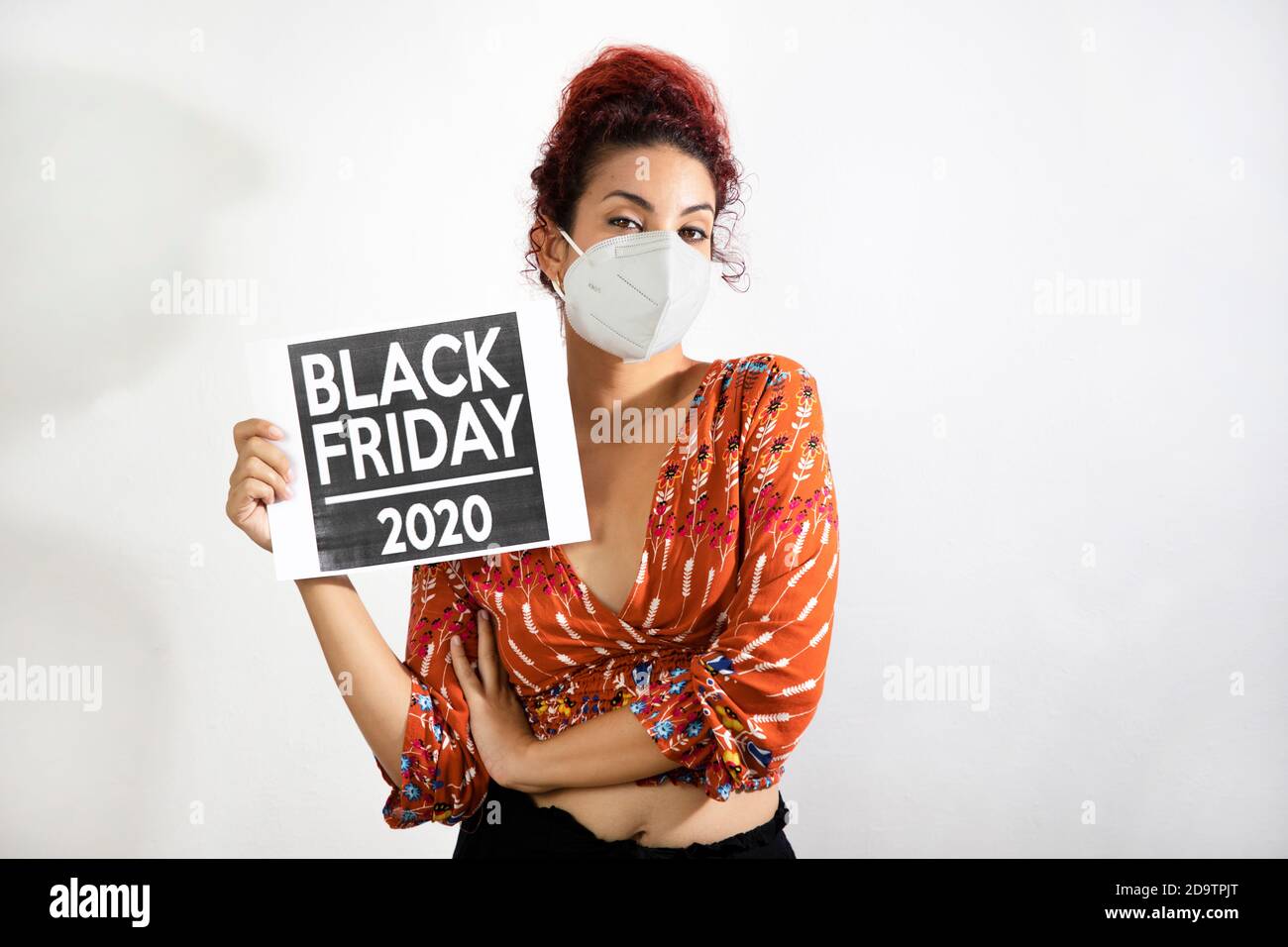 Mädchen mit Maske hält ein schwarzes freitag 2020 Poster mit ihrer Hand. Junge Frau mit weißer Haut und lockigen roten Haaren in bunt gemusterter Kleidung gekleidet Stockfoto